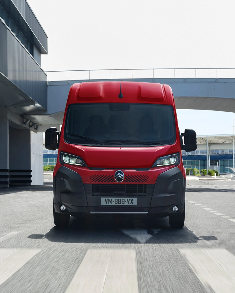 Vooraanzicht van een rode Citroën Jumper-bestelwagen die door een moderne stedelijke omgeving rijdt, waarbij de nadruk ligt op het robuuste ontwerp, de functionaliteit en de opvallende aanwezigheid op de weg.