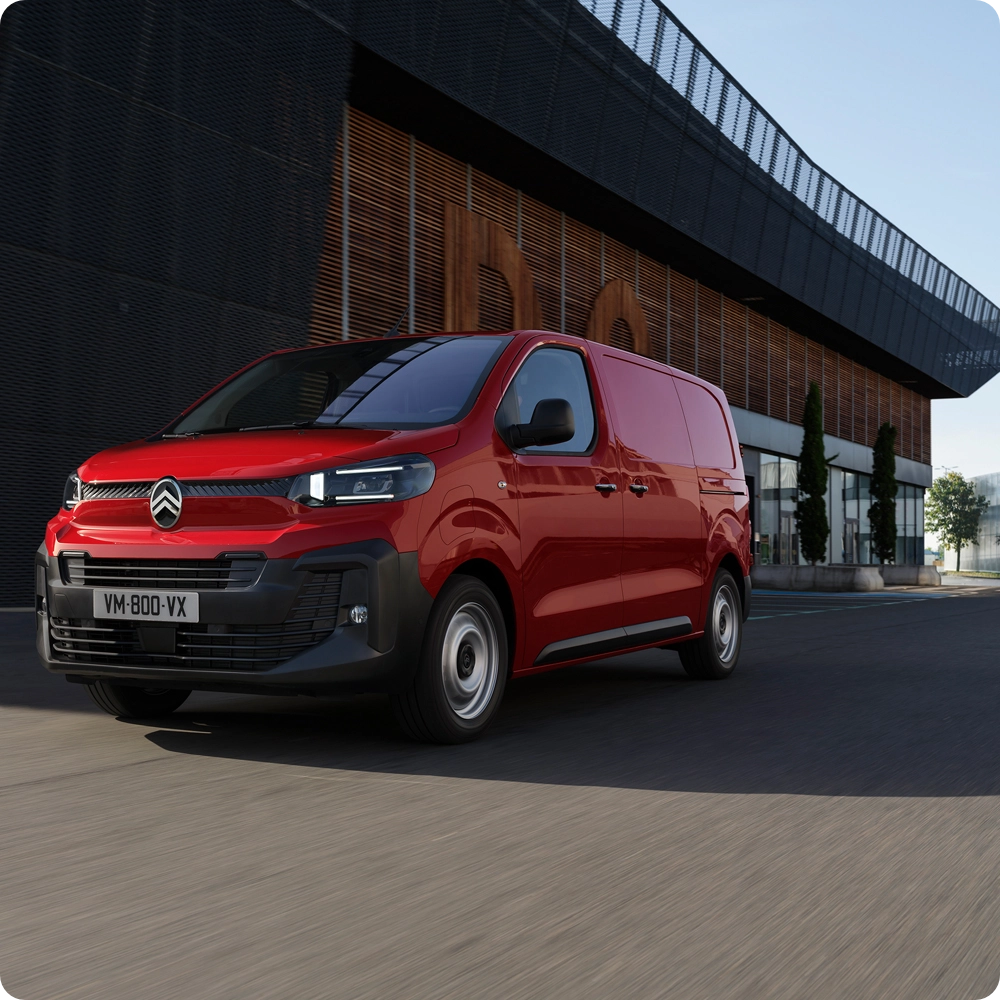 Three‑quarter front view of a red Citroën ë-Jumpy driving in an urban environment, showing modern design, driving comfort, connectivity and confident professional presence.