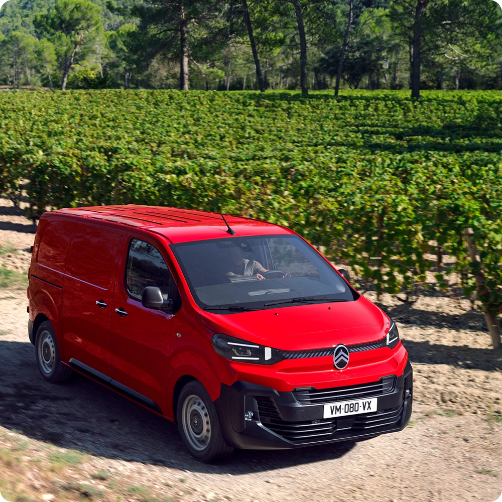 Three‑quarter front exterior view of a red Citroën ë-Jumpy driving in an urban environment, showing safety features, driving comfort and confident professional presence.