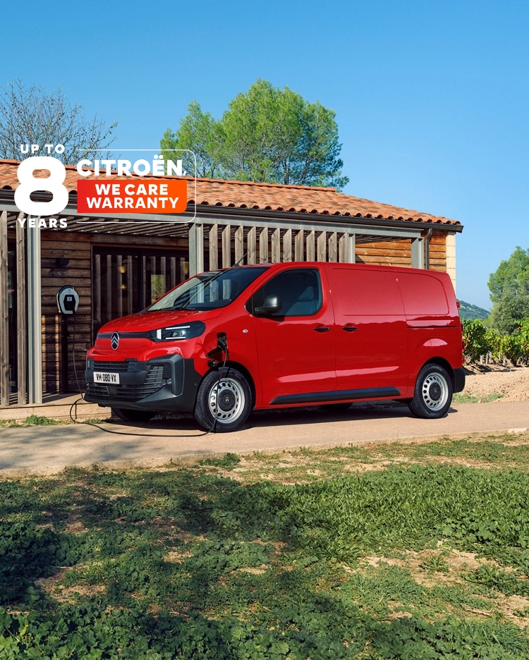 Side three‑quarter exterior view of a red Citroën ë‑Jumpy charging in front of a building, showing modern electric design, everyday practicality, connectivity and confident professional presence.