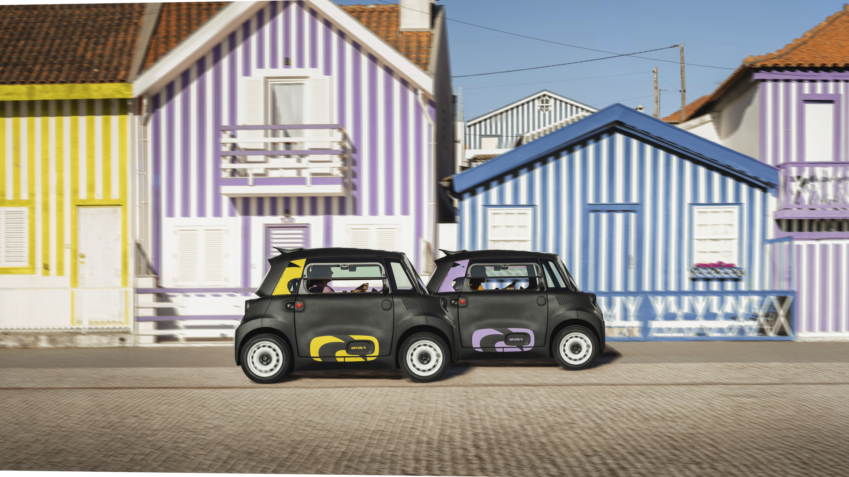 Straataanzicht naast elkaar van de Citroën Ami x Rip Curl elektrische wagen zonder rijbewijs, dubbele Citroën Ami‑stijl voor een stedelijke elektrische wagen