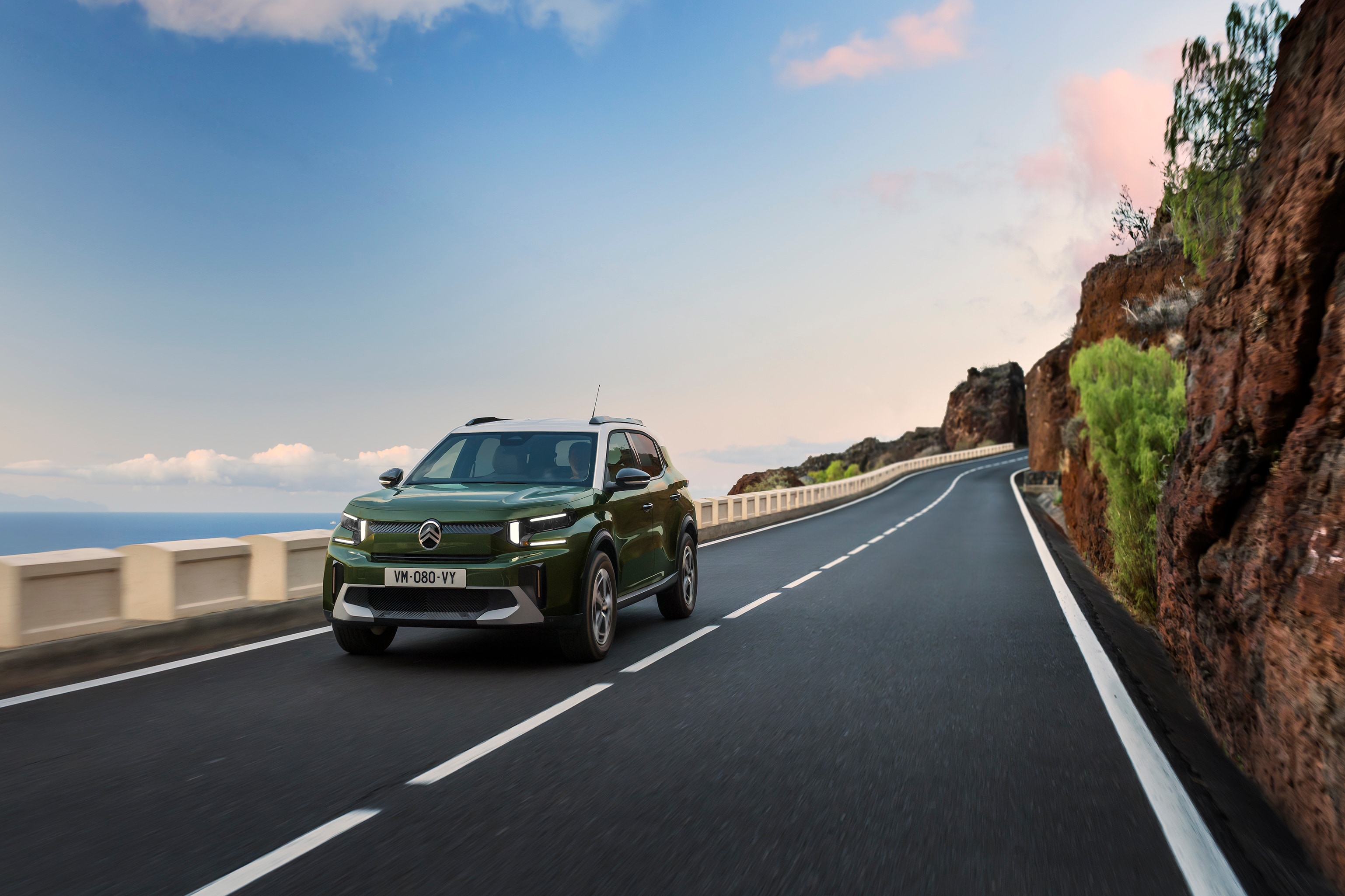 Voorste driekwart rijbeeld van de Citroën ë-C3 compacte SUV op een schilderachtige weg die wendbare, zelfverzekerde en betaalbare dagelijkse mobiliteit benadrukt.