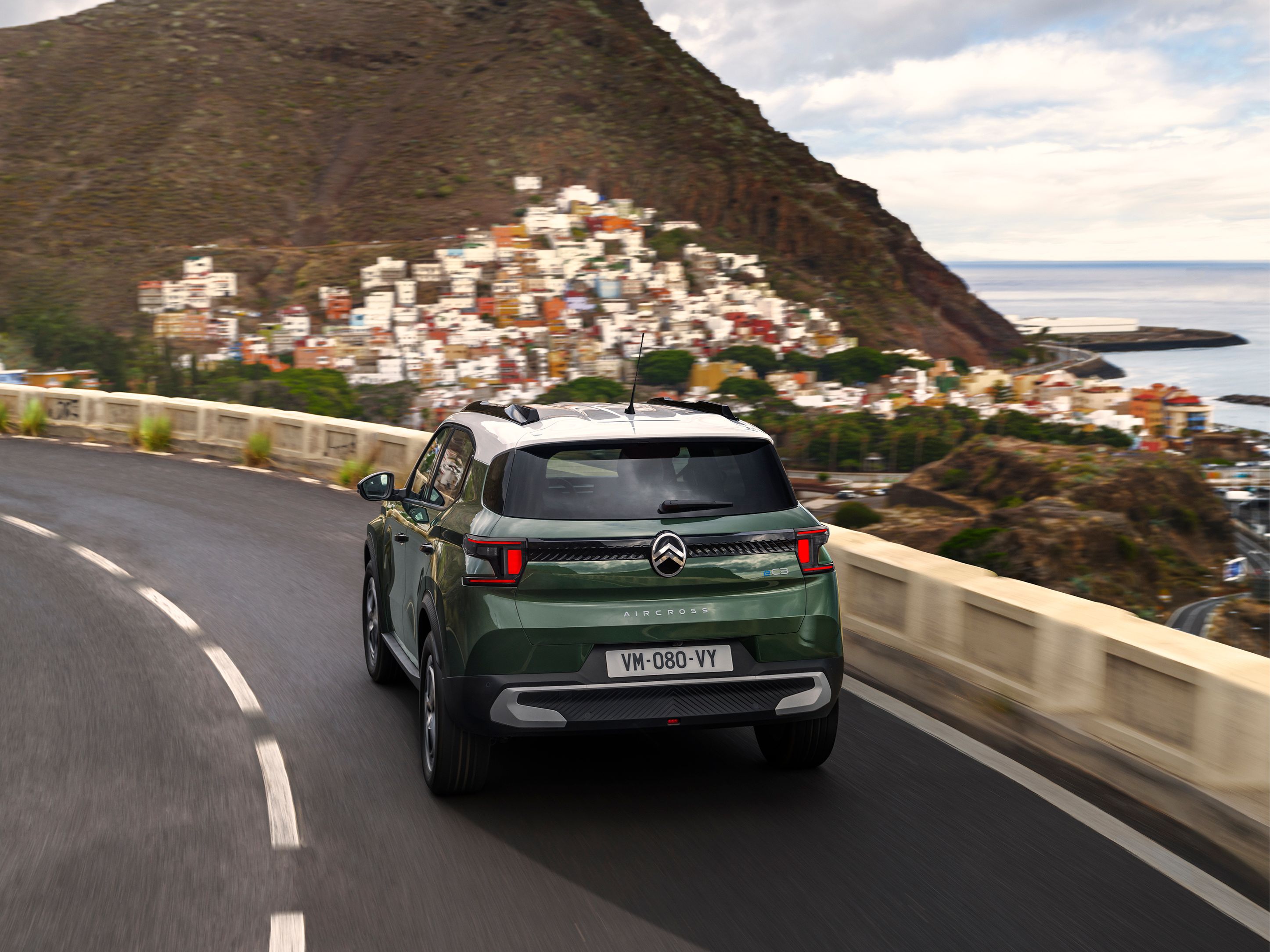 Achterste driekwart rijbeeld van de Citroën ë-C3 Aircross SUV op een kustweg voor zelfverzekerde, soepele en betaalbare dagelijkse mobiliteit.